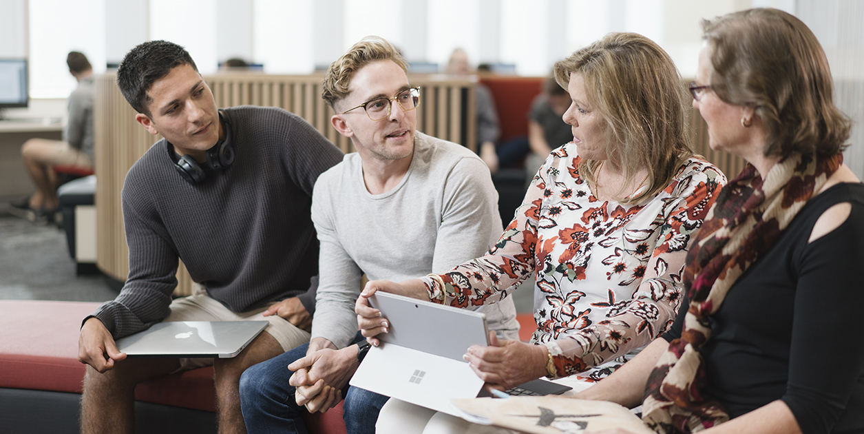 Four adults sitting on a bench in a library area, holding laptops and computers