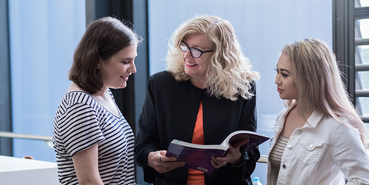 Two students stand alongside a smiling academic who holds an open book