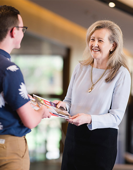 An older, finely and professionally dressed woman hands a young student some booklets 