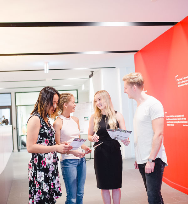 Four students walk through a well-lit hallway, looking at brochures and talking.