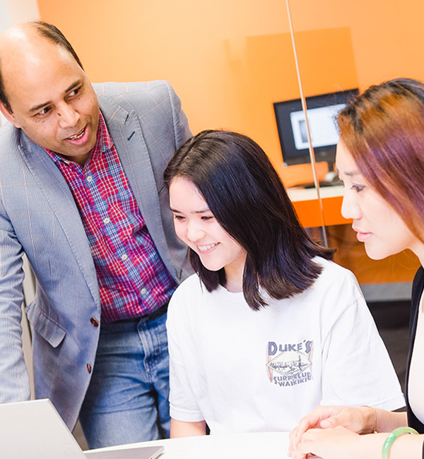 Two ACU students sit at a laptop while an instructor stands and talks to them